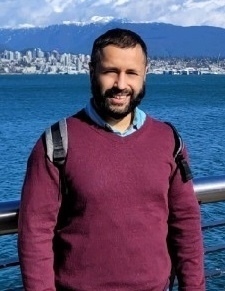 Man with a dark beard, wearing a maroon sweater and backpack, smiles with a cityscape and mountains across the water in the background.